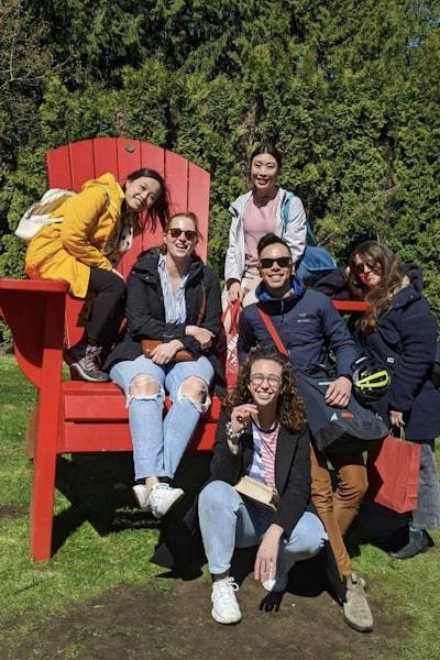 A group of young people smiling, sitting in the sunshine. Backround is leafy and green and the grass is green. Three people are sitting on a giant red wooden chair, while two are leaning against it and one is sitting in front of it. 