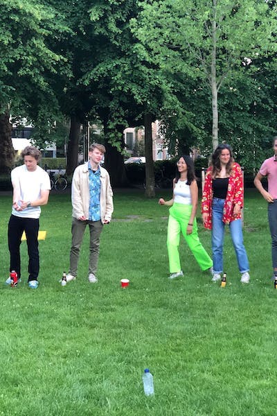 Group of young people standing in a park playing games involving throwing bottles. Two young men, two young women, wearing t-shirts and jackets, colorful trousers and patterned shirts. 