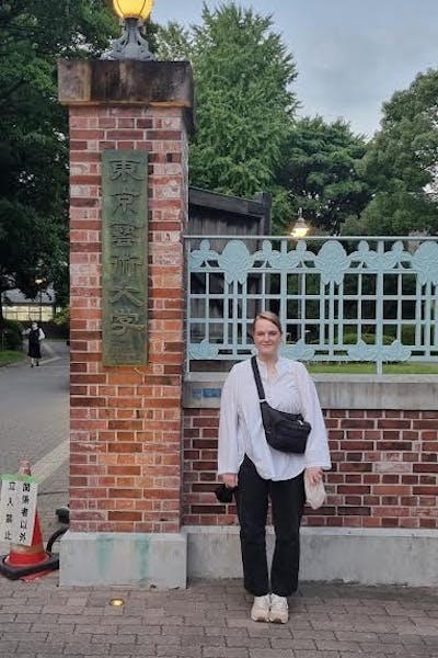 Young woman standing in front of a red brick fence with Japanese letters on it.  She has blond hair and is wearing a white shirt and black trousers. 