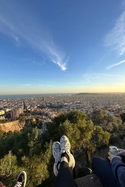 A view from a hilltop over the city of Barcelona. In the forefront there are the feet and shoes of those who took the photo. The sky is blue and the sun is about to set. 