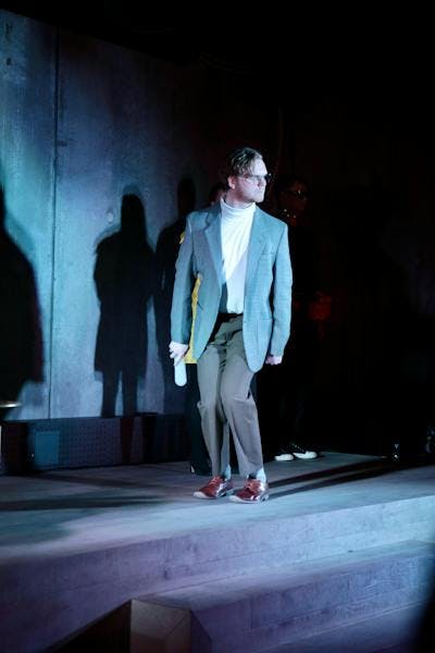 Man in a gray suit walking on stage in a theater production. He has glasses and brown hair and is also wearing a white turtle neck shirt.
