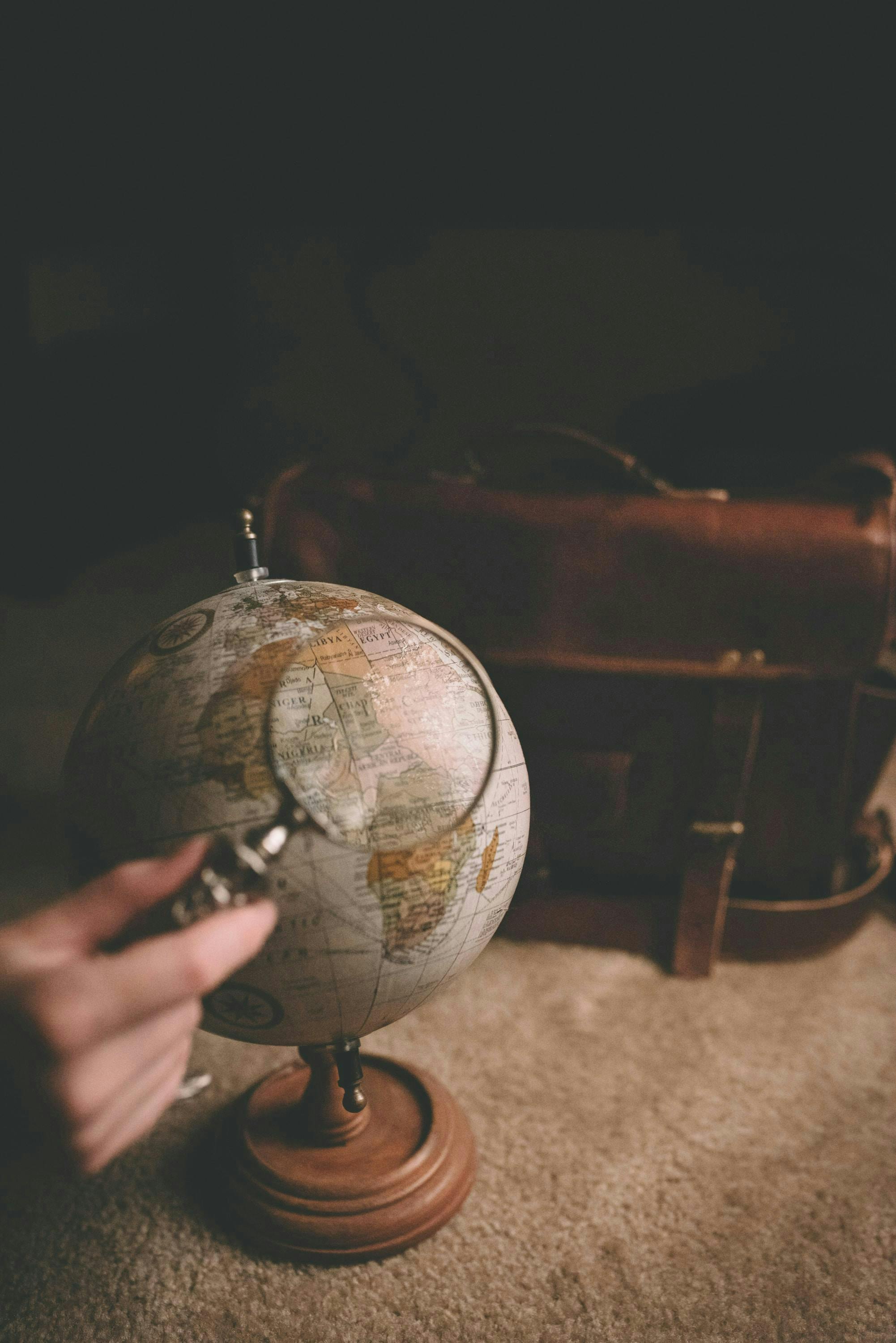Black background, dark suitcase standing on beige carpet. In front of suitcase an old fashioned globe behind a hand holding a magnifying glass
